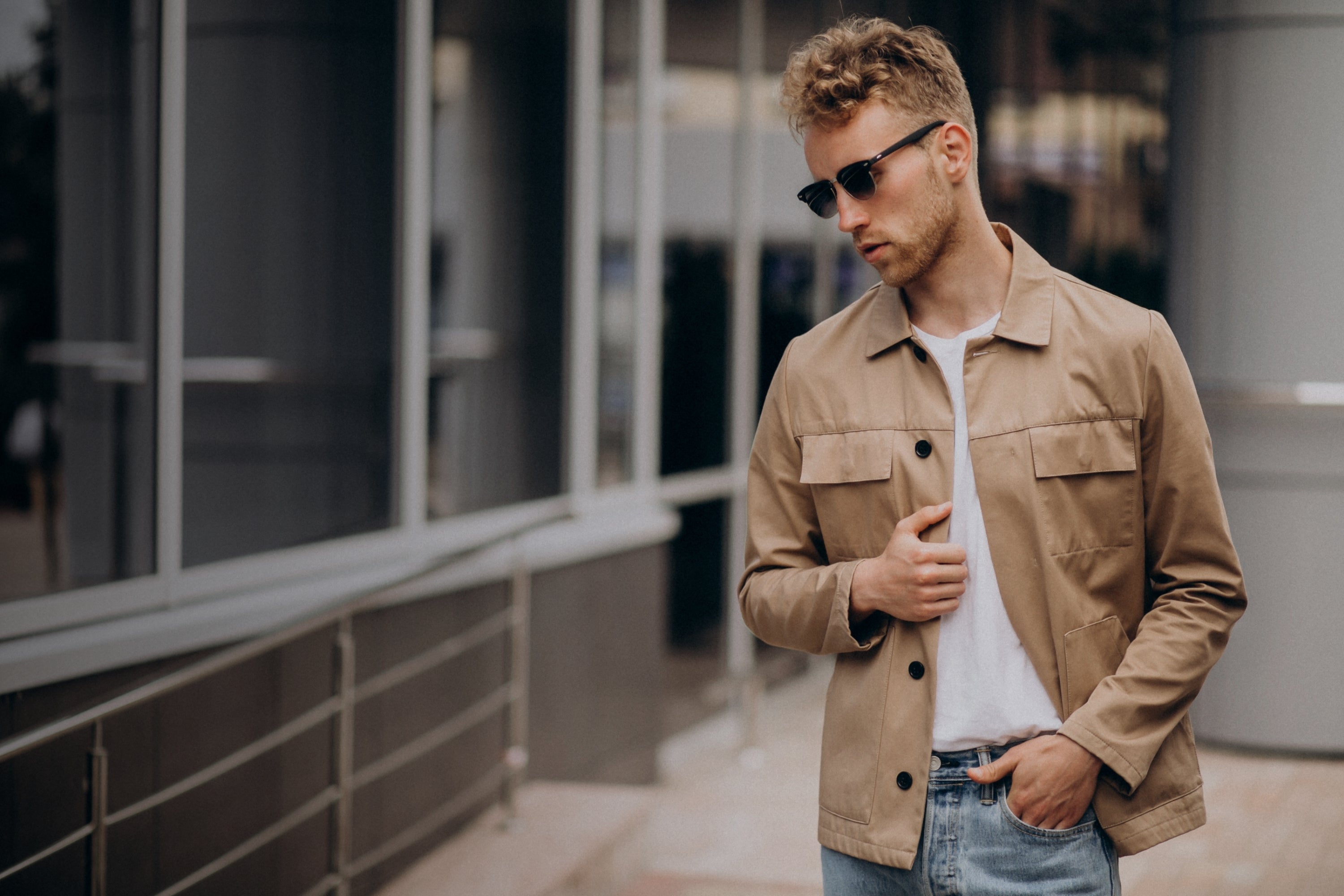 Man wearing a beige jacket and sunglasses standing outdoors.