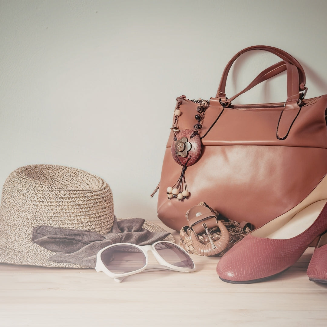 Brown handbag, pink shoes, straw hat, and sunglasses on a light wooden surface.